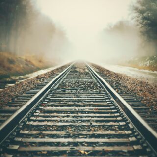 Railroad tracks disappearing into a foggy, sunlit distance surrounded by autumn trees and fallen leaves.