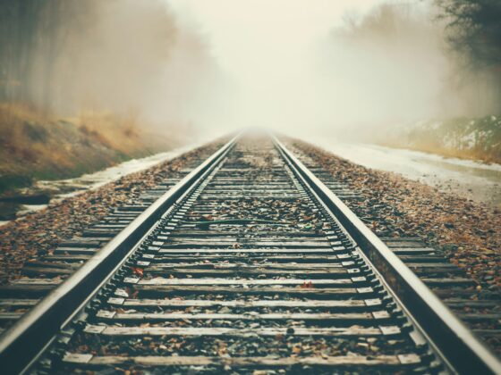 Railroad tracks disappearing into a foggy, sunlit distance surrounded by autumn trees and fallen leaves.