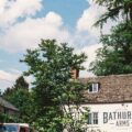Front of a white brick pub with a large Bathurst Arms sign, framed by green trees against a blue sky with clouds.