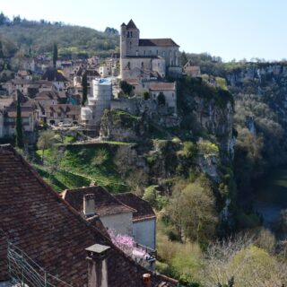 Medieval village perched on a rocky hillside above a winding river, with a tall church tower at the center.