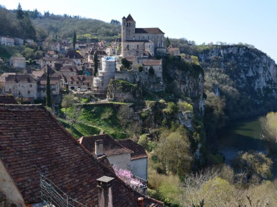 Medieval village perched on a rocky hillside above a winding river, with a tall church tower at the center.