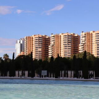 Row of tan apartment buildings behind a line of tall trees, with a pool in the foreground and blue sky above