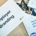 Close-up of marketing brochures on a wooden desk, with bold 'Employer Branding' text and a photo collage on one page and blue/orange design accents nearby.