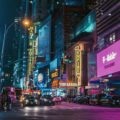 Bustling Times Square at night with neon signs, crowds on the crosswalk, and traffic in neon pink and blue reflections. Signs show theater and brand ads across tall buildings.
