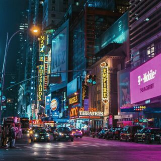 Bustling Times Square at night with neon signs, crowds on the crosswalk, and traffic in neon pink and blue reflections. Signs show theater and brand ads across tall buildings.