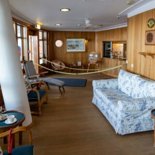 Wood-paneled boat lounge with floral sofa, wicker chairs, and a rope barrier across the room.