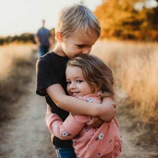 Two young children hug each other in a sunlit field with autumn trees in the background, warm golden light.