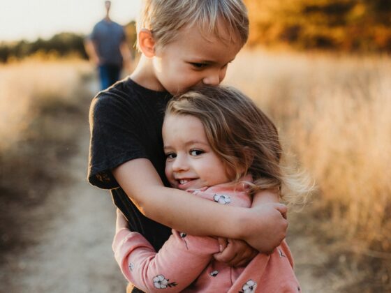 Two young children hug each other in a sunlit field with autumn trees in the background, warm golden light.