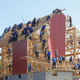 Construction workers in hats assemble a wooden roof frame on a two-story building, with red corrugated metal panels and a clear blue sky behind them.