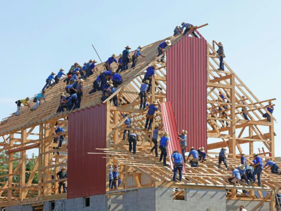 Construction workers in hats assemble a wooden roof frame on a two-story building, with red corrugated metal panels and a clear blue sky behind them.