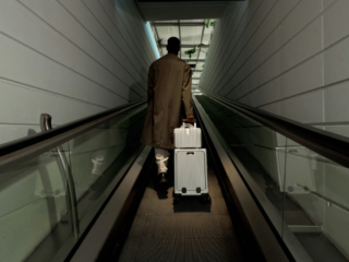 Person in a brown trench coat walks away on an airport moving walkway, pulling a silver rolling suitcase behind them.