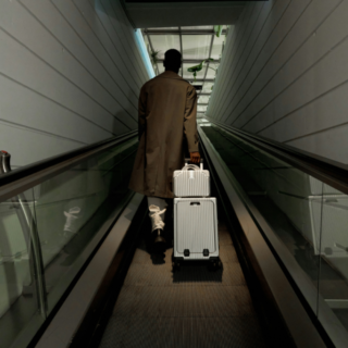 Person in a brown trench coat walks away on an airport moving walkway, pulling a silver rolling suitcase behind them.