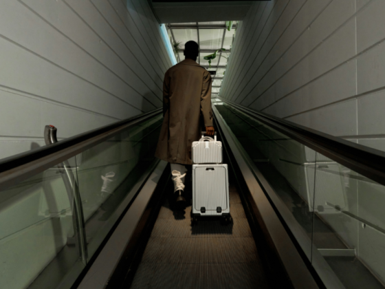 Person in a brown trench coat walks away on an airport moving walkway, pulling a silver rolling suitcase behind them.