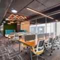 Open-plan coworking space with long wooden tables, gray and yellow ergonomic chairs, and large windows along the right wall
