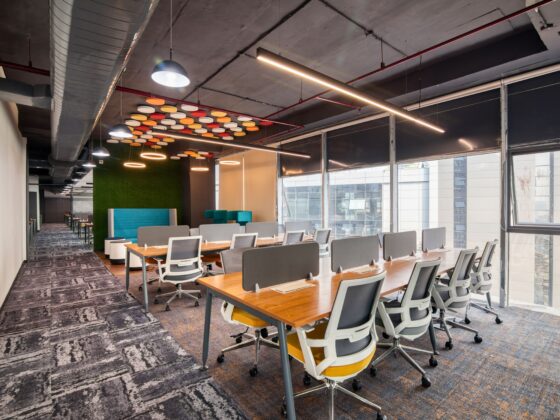 Open-plan coworking space with long wooden tables, gray and yellow ergonomic chairs, and large windows along the right wall