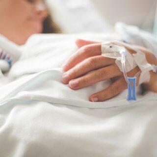 Close-up of a patient's hand with an IV catheter taped to the forearm in a hospital bed.