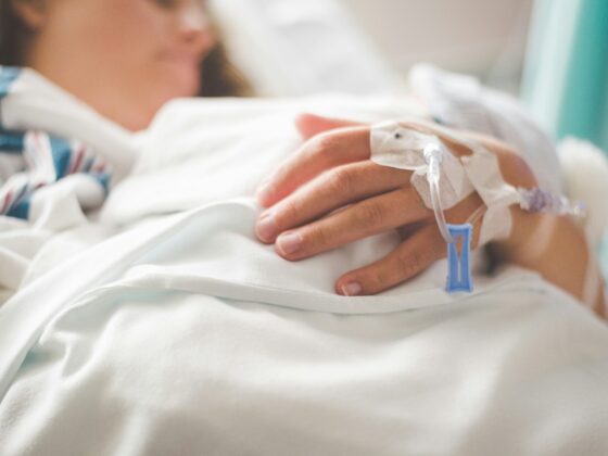 Close-up of a patient's hand with an IV catheter taped to the forearm in a hospital bed.