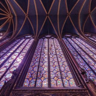 Inside a Gothic cathedral, blue starry vaulted ceiling with ribbed arches and tall colorful stained-glass windows
