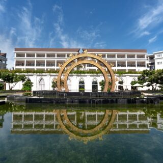 Resort courtyard with white buildings and a decorative gold circular arch sculpture over a reflecting pool, blue sky above; reflection visible in water