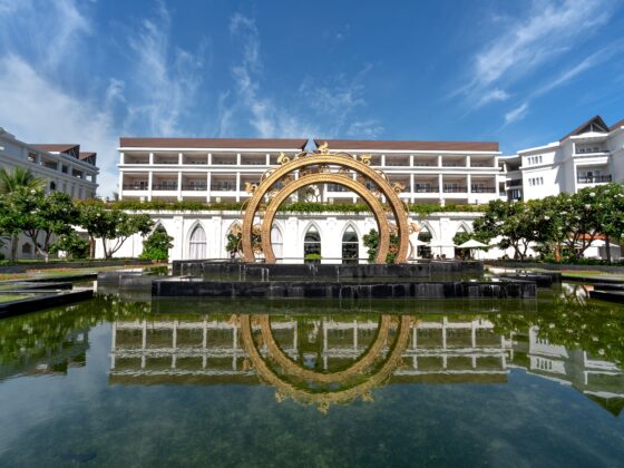 Resort courtyard with white buildings and a decorative gold circular arch sculpture over a reflecting pool, blue sky above; reflection visible in water