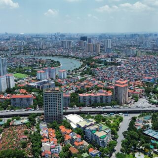 Aerial view of a sprawling city with high-rise towers, dense housing, and winding canals; a large elevated highway runs across the bottom edge.