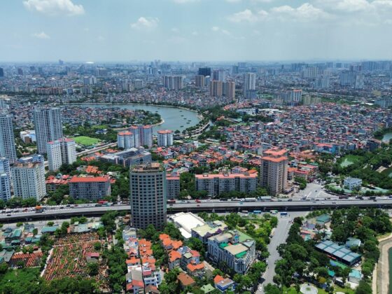 Aerial view of a sprawling city with high-rise towers, dense housing, and winding canals; a large elevated highway runs across the bottom edge.