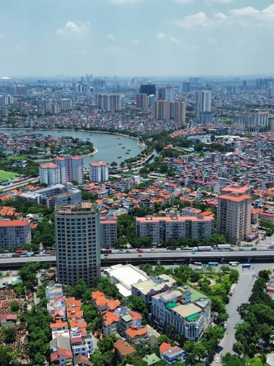 Aerial view of a sprawling city with high-rise towers, dense housing, and winding canals; a large elevated highway runs across the bottom edge.