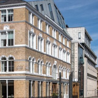 Brick building with arched windows and white trim on a sunny city street, with a modern glass-and-concrete building beside it.