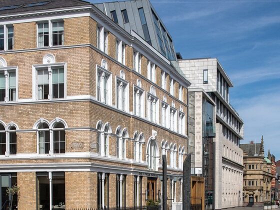 Brick building with arched windows and white trim on a sunny city street, with a modern glass-and-concrete building beside it.