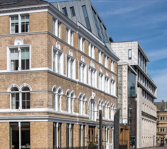 Brick building with arched windows and white trim on a sunny city street, with a modern glass-and-concrete building beside it.