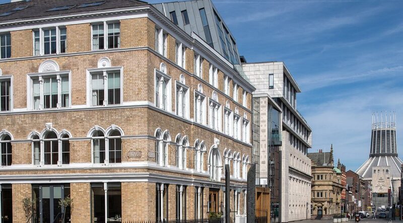 Brick building with arched windows and white trim on a sunny city street, with a modern glass-and-concrete building beside it.