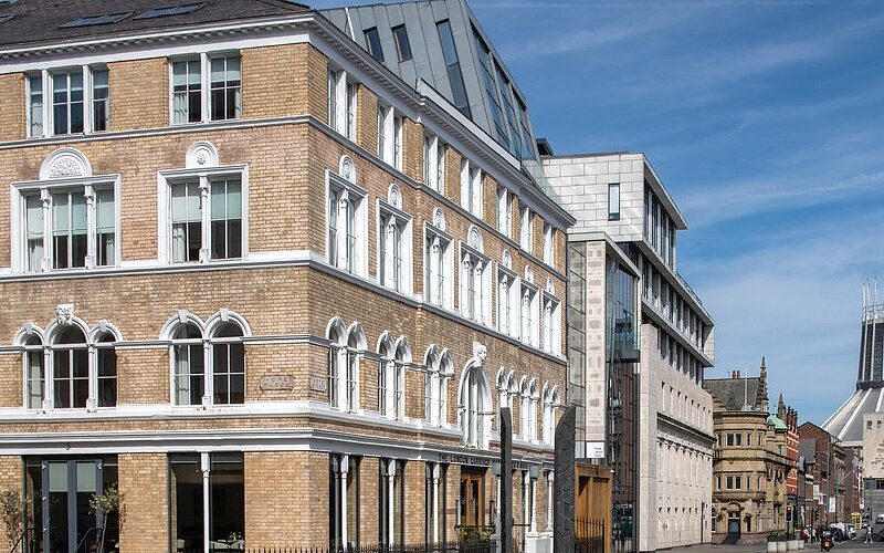 Brick building with arched windows and white trim on a sunny city street, with a modern glass-and-concrete building beside it.