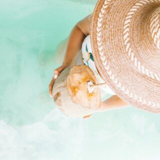 Person viewed from above, wearing a large woven straw hat and holding a coconut drink at the beach with turquoise water in the background.