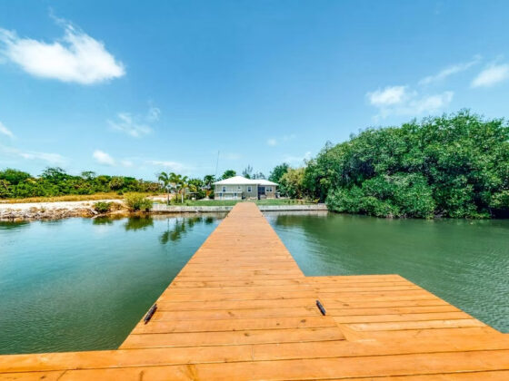 Long wooden dock extends over calm water toward a waterfront house under a bright blue sky, with trees on the right.