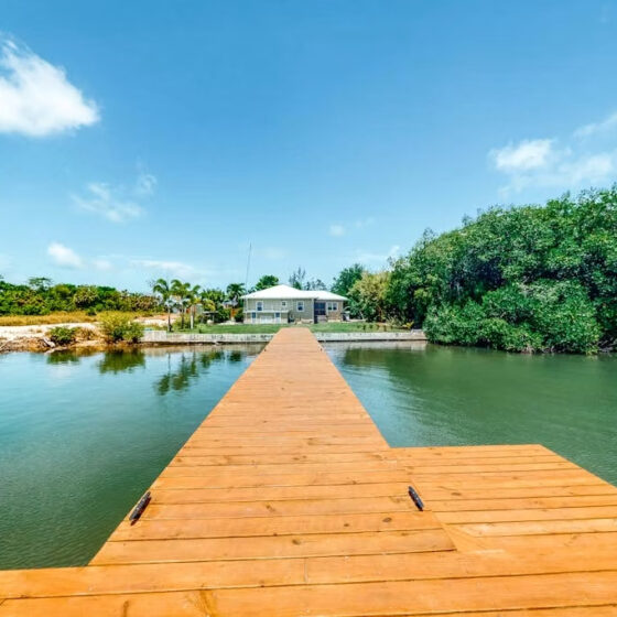 Long wooden dock extends over calm water toward a waterfront house under a bright blue sky, with trees on the right.