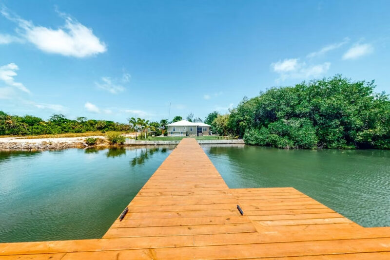 Long wooden dock extends over calm water toward a waterfront house under a bright blue sky, with trees on the right.