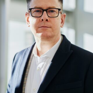 Portrait of a man with dark hair and black-r framed glasses, wearing a white shirt and navy blazer, standing in a bright indoor space with soft natural light.