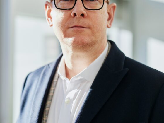 Portrait of a man with dark hair and black-r framed glasses, wearing a white shirt and navy blazer, standing in a bright indoor space with soft natural light.