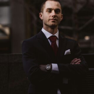 Confident man in a dark suit and red tie, arms crossed, standing outdoors at dusk, looking at the camera.