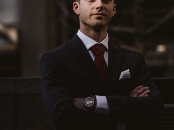 Confident man in a dark suit and red tie, arms crossed, standing outdoors at dusk, looking at the camera.