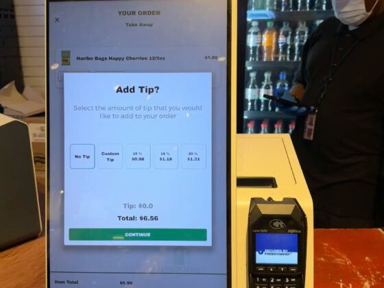 Touchscreen self-checkout kiosk showing an order summary and tip options, with a person in a mask nearby at the counter.
