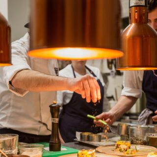 Chef plating elegant dishes under warm copper pendant lights, sprinkling garnish onto plates in a busy kitchen.
