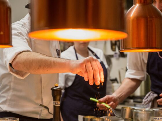 Chef plating elegant dishes under warm copper pendant lights, sprinkling garnish onto plates in a busy kitchen.