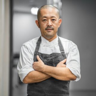 Male chef with a shaved head wearing a white jacket and dark apron, arms crossed in a kitchen corridor.