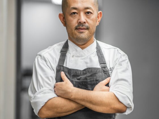 Male chef with a shaved head wearing a white jacket and dark apron, arms crossed in a kitchen corridor.