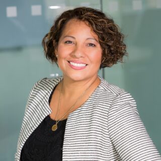 Smiling woman with short curly hair wearing a striped blazer and black lace top in an office setting with glass walls behind her.