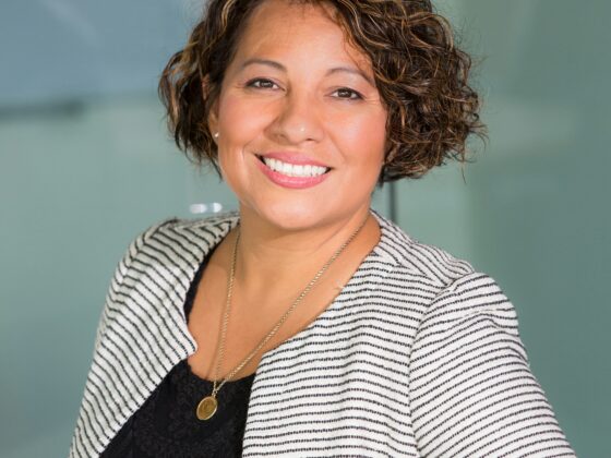 Smiling woman with short curly hair wearing a striped blazer and black lace top in an office setting with glass walls behind her.