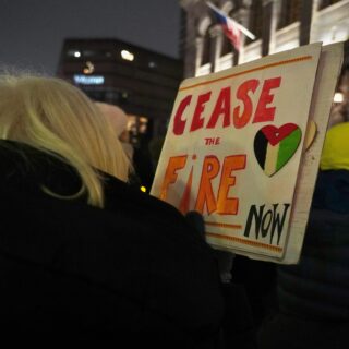 Protester seen from behind at night, holding a sign that reads 'PLEASE FIRE NOW' with a heart graphic and a city street in the background.