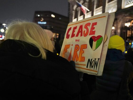Protester seen from behind at night, holding a sign that reads 'PLEASE FIRE NOW' with a heart graphic and a city street in the background.