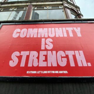 Red storefront banner with large white text reading 'COMMUNITY IS STRENGTH' and smaller line 'Be strong. Let's look out for one another.' A small 'ALLIED' label sits at the top edge; an old brick building is visible above the sign.